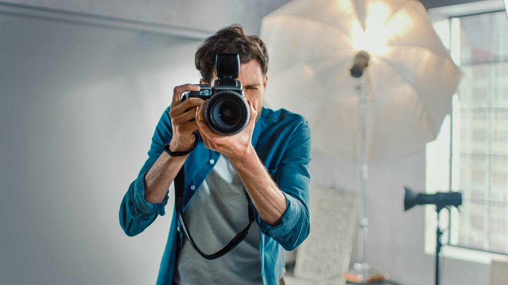 Un fotografo in camicia blu scatta una foto in uno studio con illuminazione intensa e attrezzatura visibile sullo sfondo.