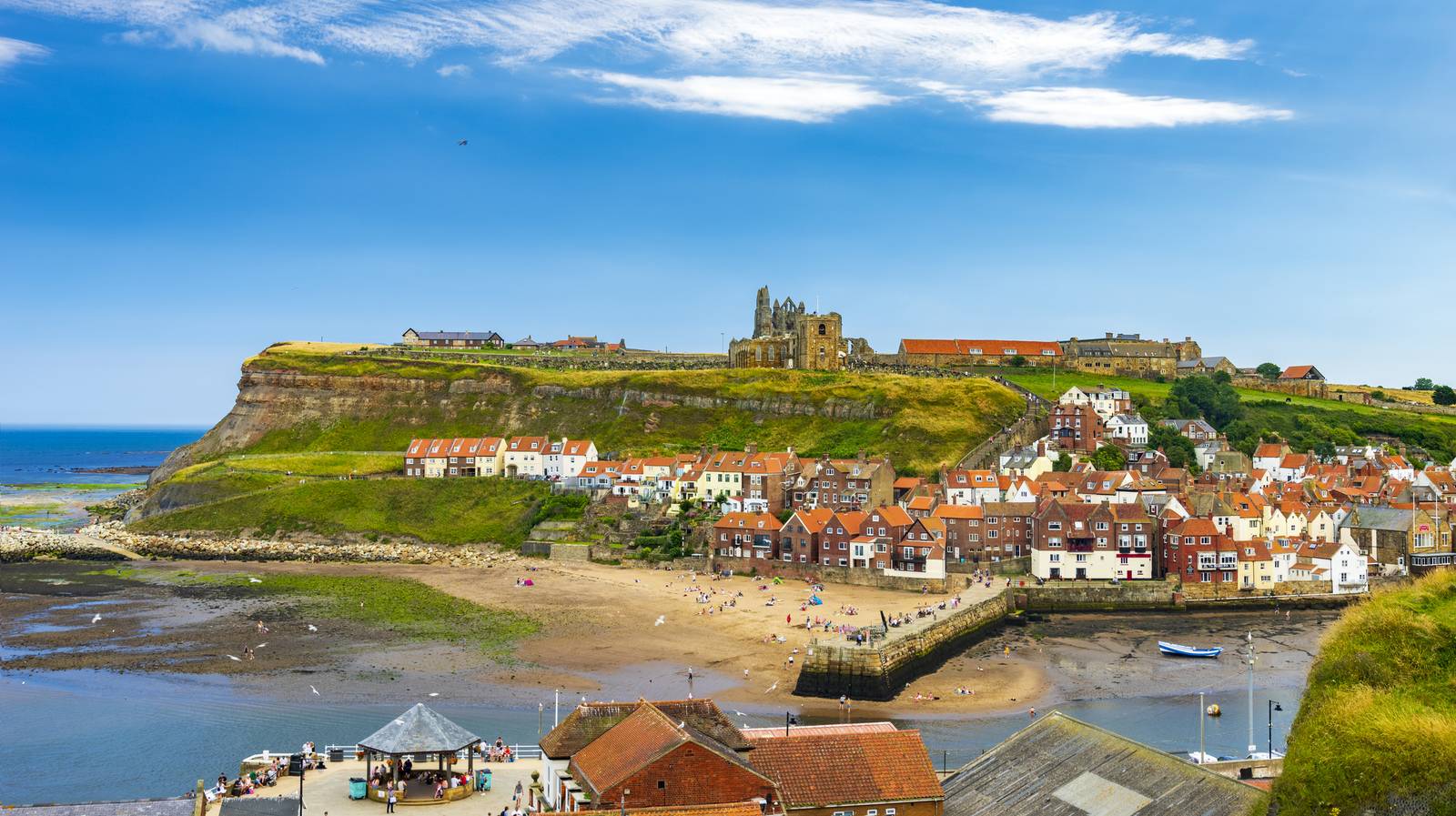 Coastal town with red-roofed houses, a sandy beach, and a hilltop ruin under a blue sky.