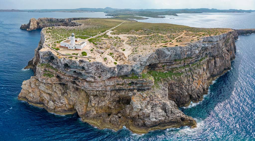 cami-de-cavalls Aerial view of a rugged coastal cliff with a lighthouse atop, surrounded by blue sea and distant land in the background.