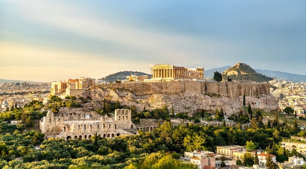 View of the Acropolis in Athens, Greece, featuring the Parthenon and surrounding ruins, with a sunny sky and the cityscape below.