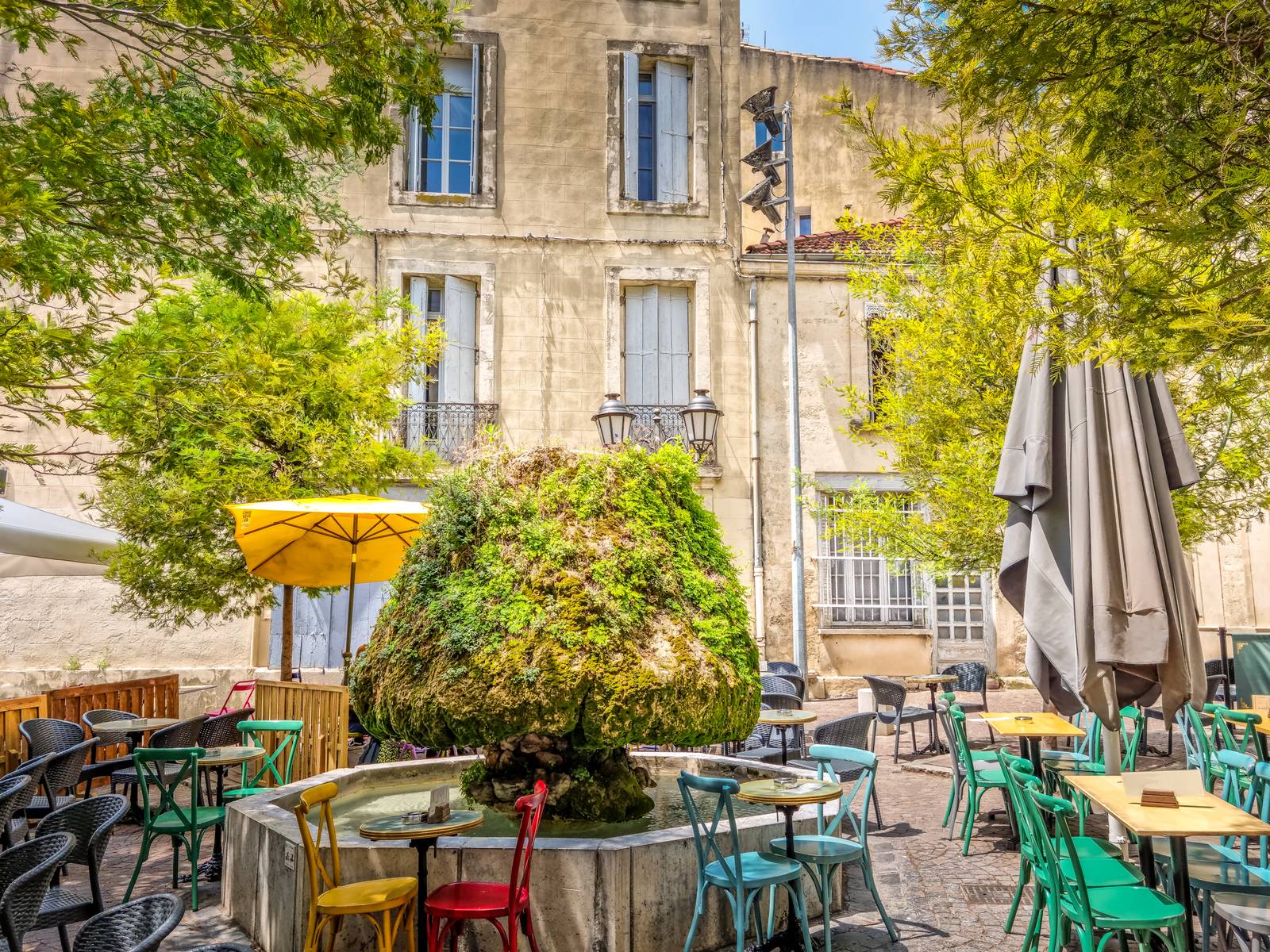Outdoor cafe in Montpellier with colorful chairs around a tree-covered fountain, surrounded by historic stone buildings and greenery.