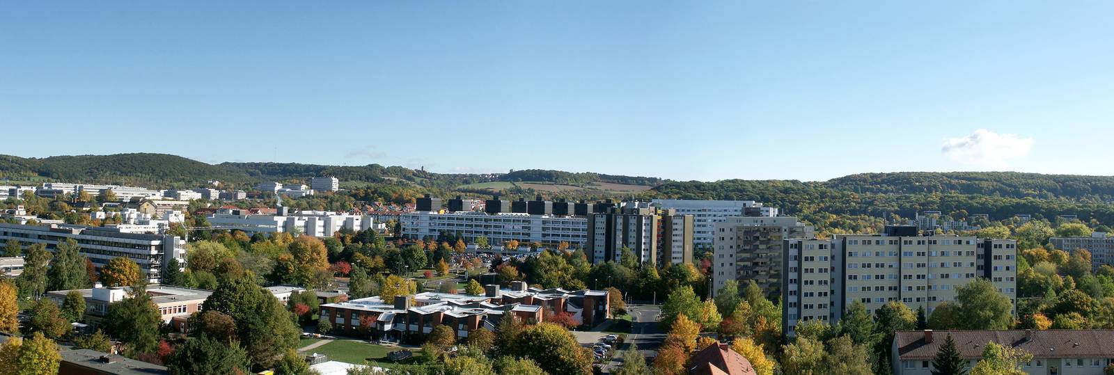 Panoramablick auf eine Stadtlandschaft mit mittelhohen Gebäuden, umgeben von üppigem Grün und Hügeln unter klarem blauem Himmel