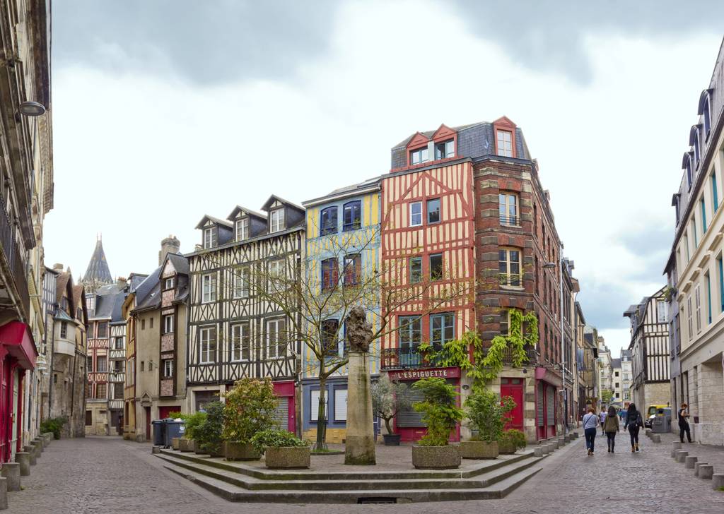 Des maisons à colombages aux couleurs vives bordent une charmante rue pavée dans une ville historique européenne, où quelques passants se promènent sous un ciel nuageux.