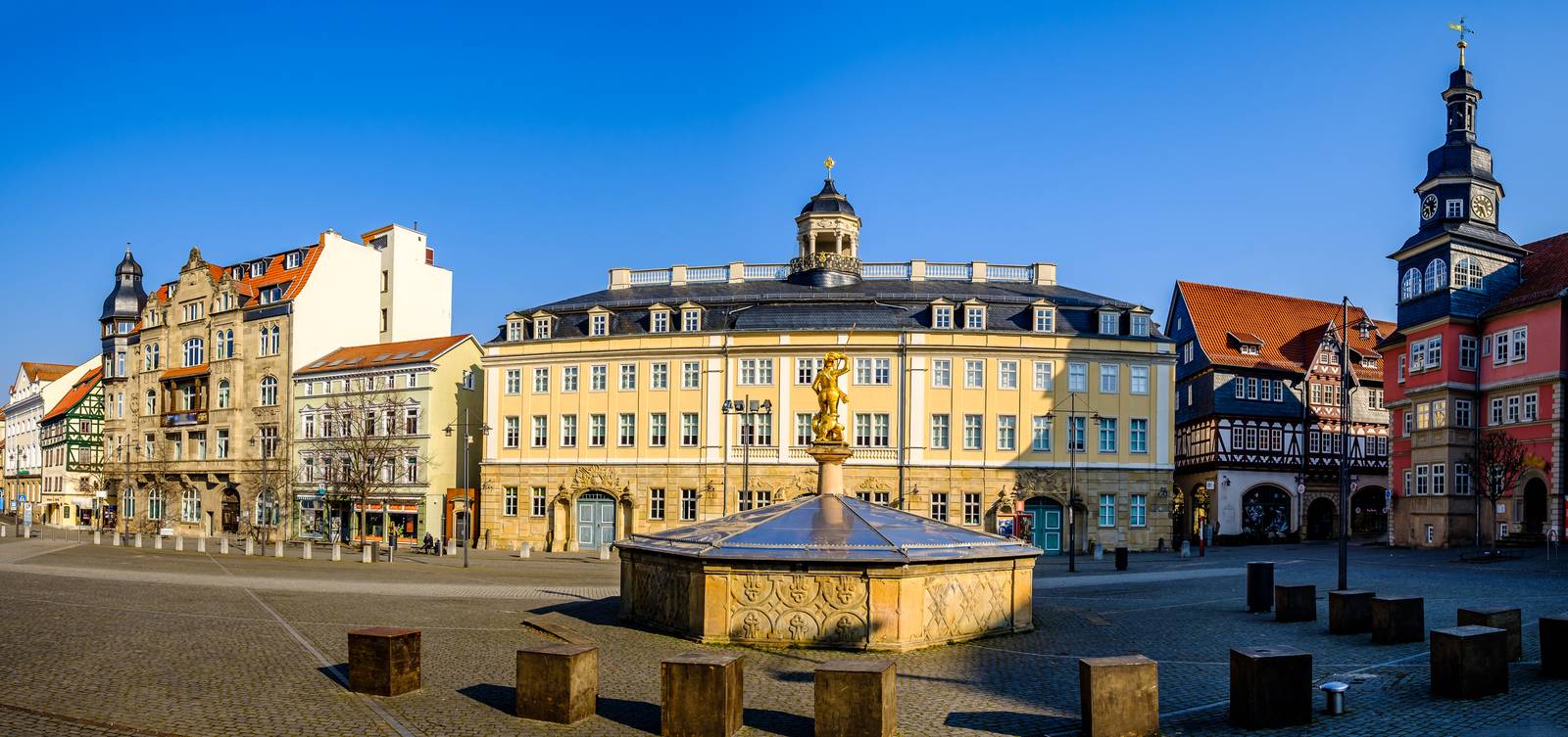 Panoramablick auf einen historischen europäischen Platz mit kunstvoll verzierten Gebäuden, einem zentralen Brunnen und klarem blauen Himmel