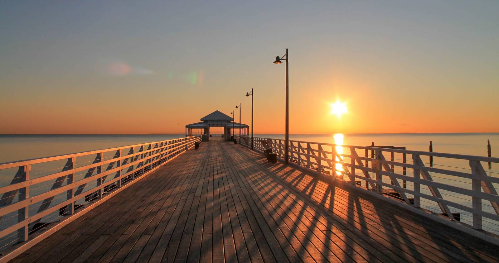 Sunrise over a wooden pier extending into calm water of Sandagate, with a gazebo at the end and warm sunlight casting long shadows.