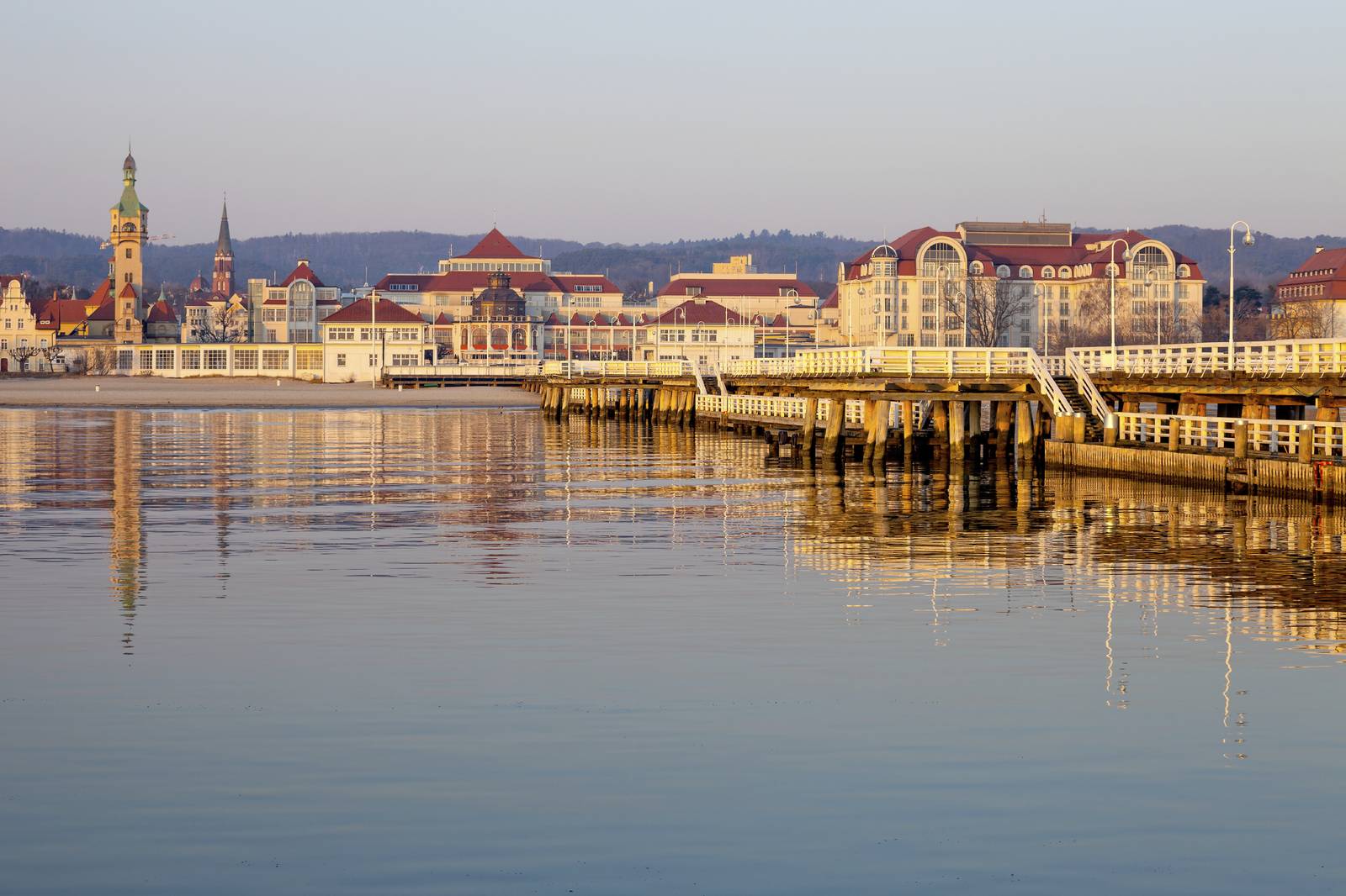 A calm waterfront scene with a pier, historic buildings, and a church tower in the background under a clear sky.