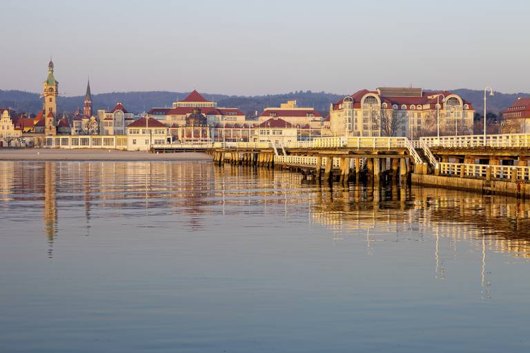 A calm waterfront scene with a pier, historic buildings, and a church tower in the background under a clear sky.