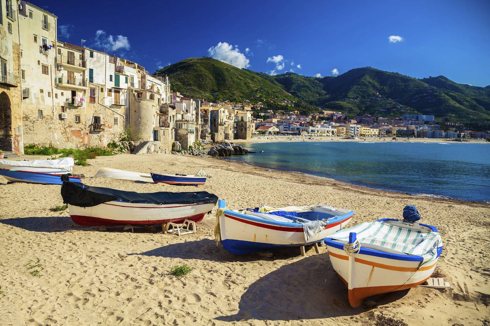 Colorful boats on a sandy beach with a coastal town and green hills in the background under a clear blue sky.