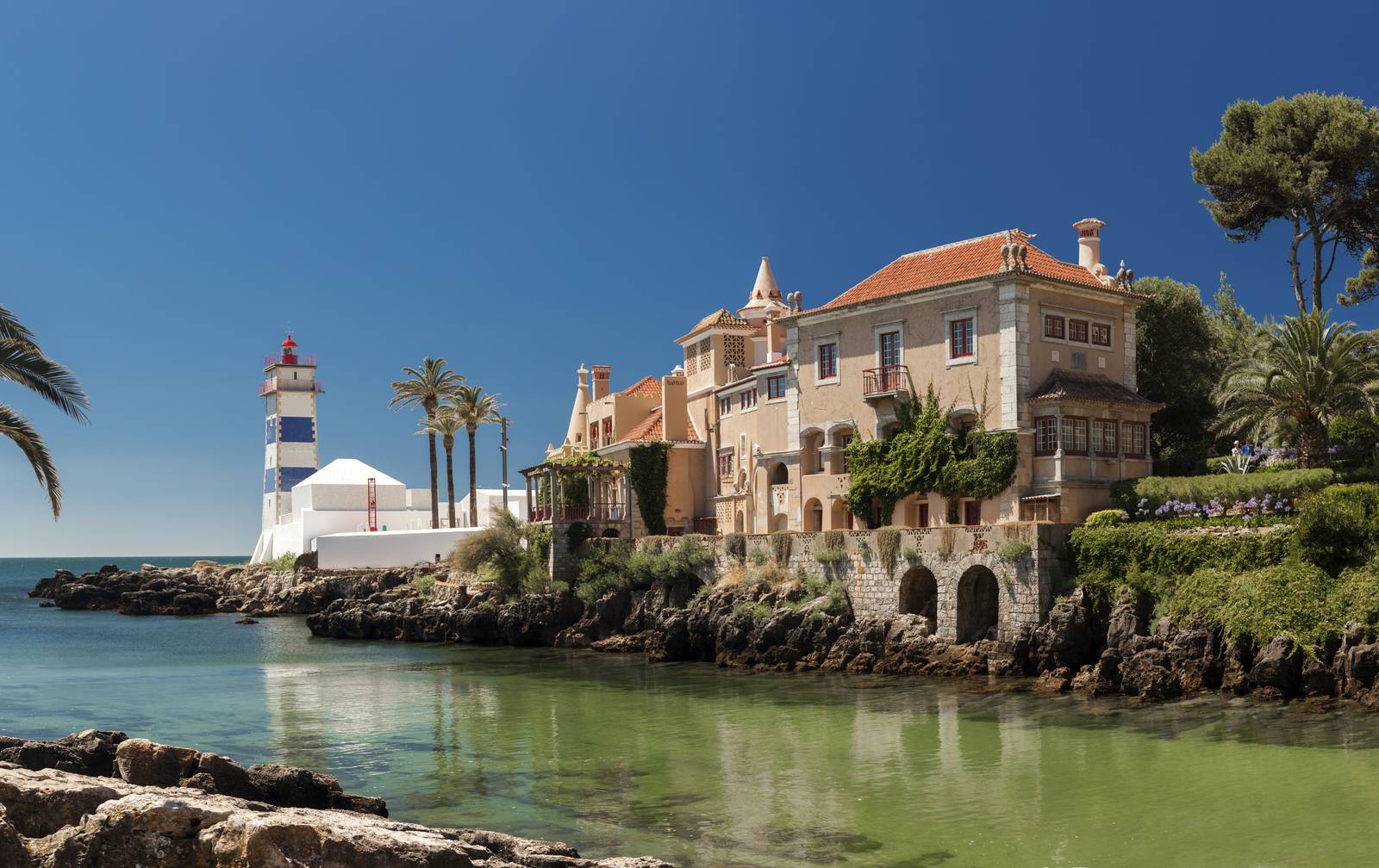 Coastal scene in Cascais with a historic building and lighthouse by calm, clear water. Palm trees and rocky shoreline under a clear blue sky.