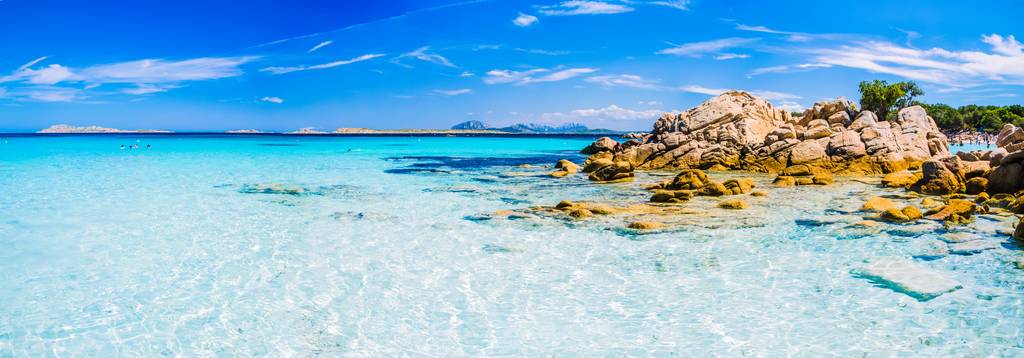 Ein atemberaubender Strand mit kristallklarem türkisfarbenem Wasser, felsiger Küste und strahlend blauem Himmel mit zarten Wolken am Horizont.