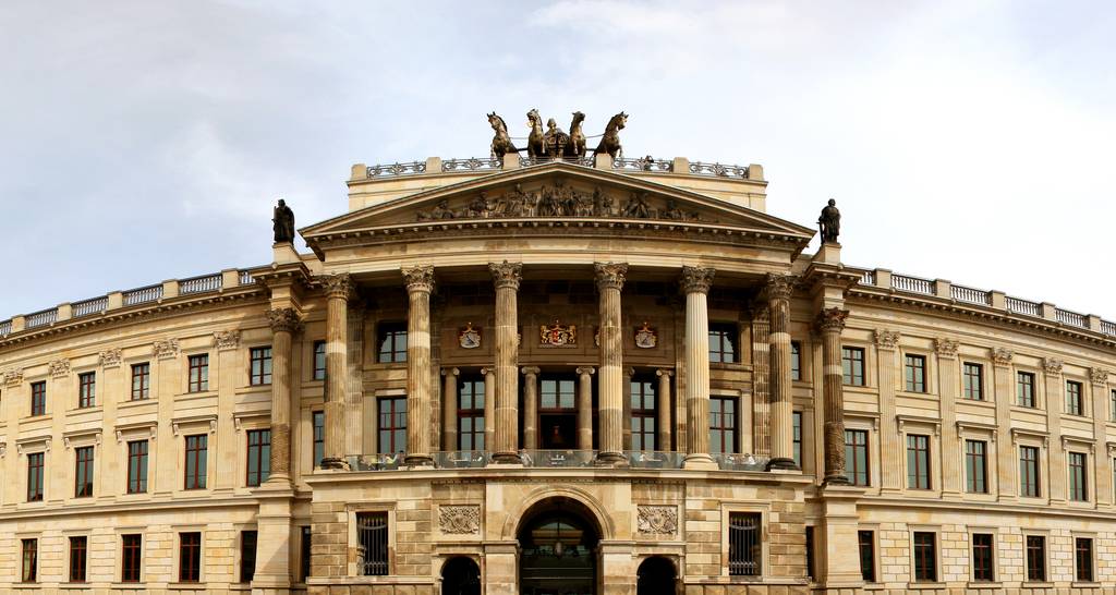 Ein prächtiges neoklassizistisches Gebäude mit kunstvoll verzierten Säulen, Statuen auf dem Dach und einem zentralen Bogeneingang, vor einem wolkenverhangenen Himmel