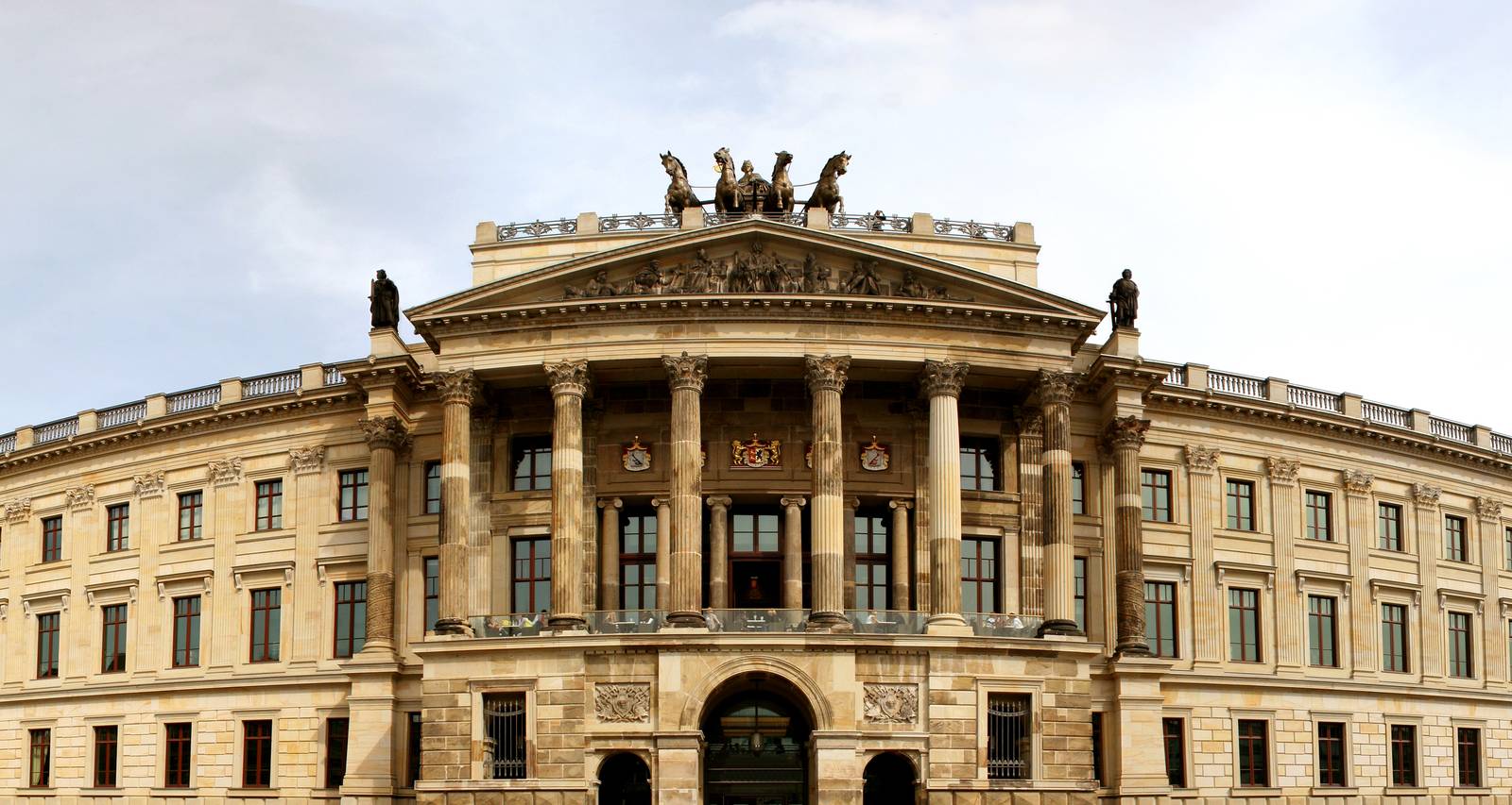 Ein prächtiges neoklassizistisches Gebäude mit kunstvoll verzierten Säulen, Statuen auf dem Dach und einem zentralen Bogeneingang, vor einem wolkenverhangenen Himmel