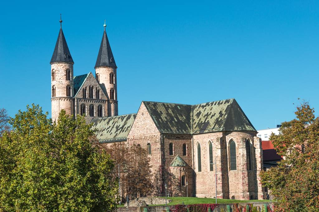 Historische Backsteinkirche mit zwei hohen, spitzen Türmen und grünem Dach, umgeben von Bäumen unter klarem, blauem Himmel