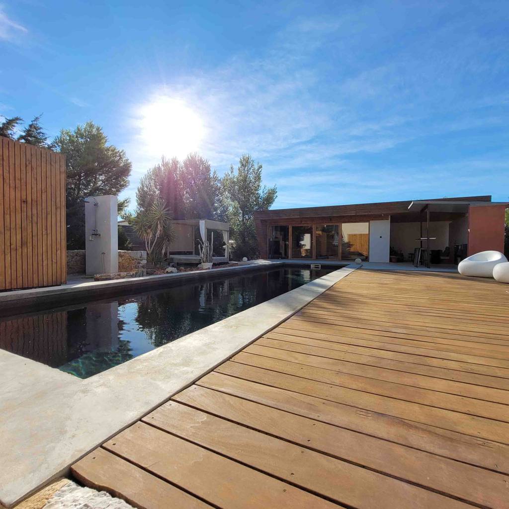 Moderne Terrasse mit Holzdeck neben einem rechteckigen Pool, umgeben von Bäumen und einem Gebäude, unter strahlender Sonne und klarem Himmel.