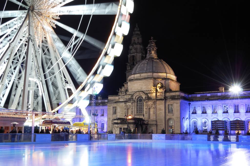 Illuminated Ferris wheel and historic building at night, with a glowing ice skating rink in the foreground, creating a festive atmosphere.