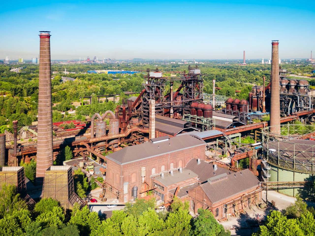 Luftaufnahme eines großen Industriekomplexes mit hohen Schornsteinen und rostenden Strukturen, umgeben von üppig grünen Bäumen unter klarem, blauem Himmel