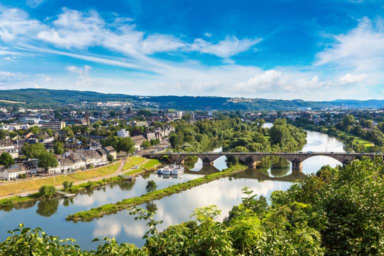 Ein malerischer Blick auf einen Fluss, der sich durch eine Stadt mit einer Steinbrücke schlängelt, umgeben von üppigem Grün und Hügeln unter einem blauen Himmel.