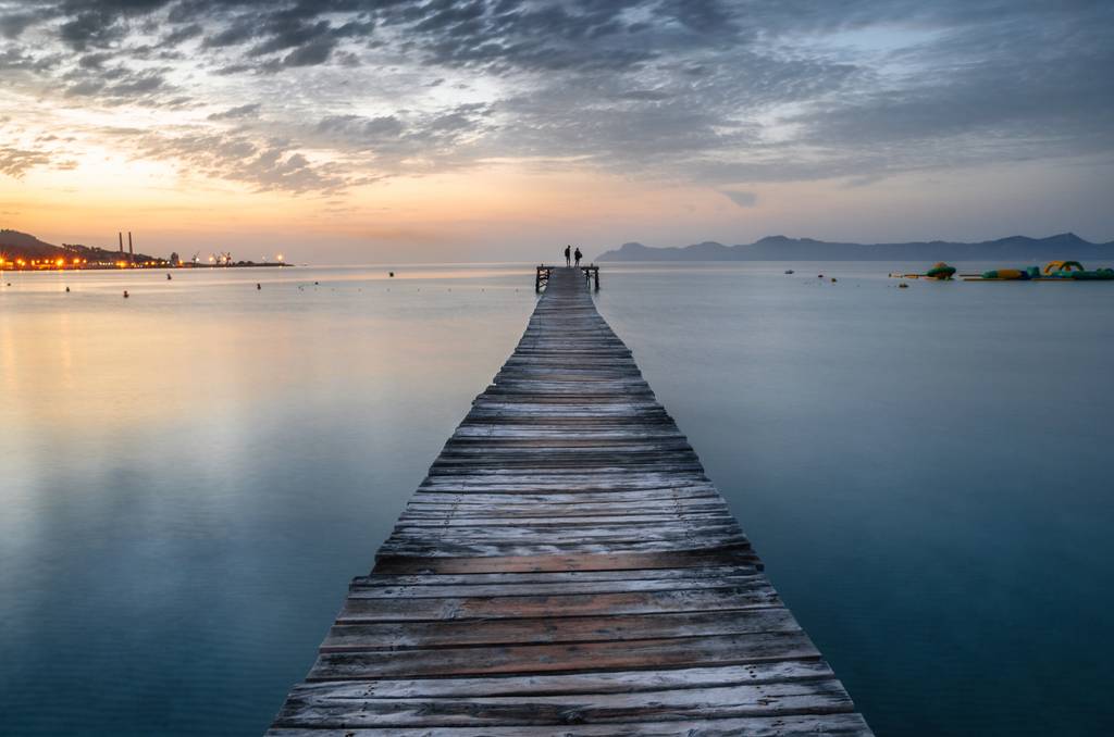 Langer Holzsteg, der bei Sonnenuntergang ins ruhige Meer führt, zwei Personen am Ende stehend, mit entfernten Lichtern und Bergen im Hintergrund.