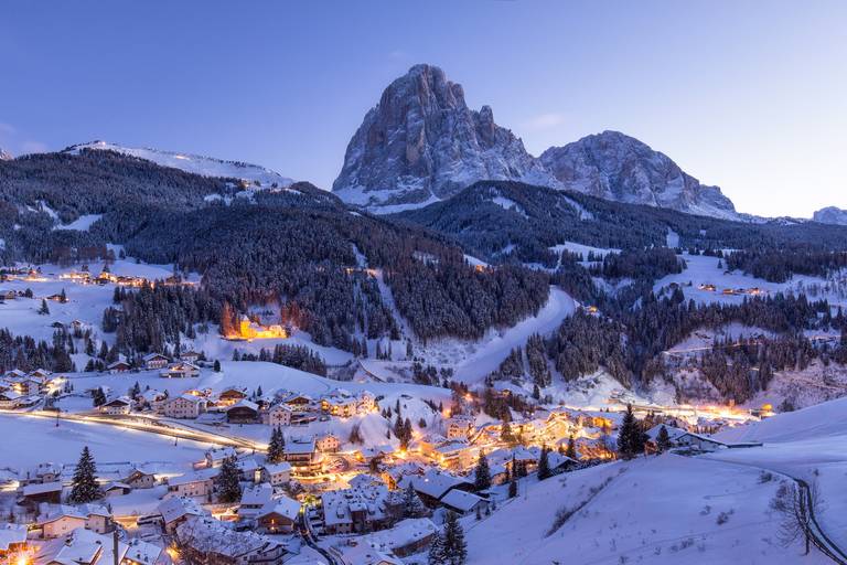 Verschneites Alpen-Dorf mit leuchtenden Lichtern in der Dämmerung, eingebettet an bewaldeten Hängen unter einem imposanten felsigen Berggipfel.