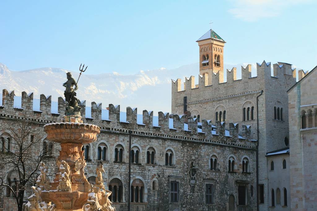 Historisches Steingebäude mit Zinnen und einem Uhrturm. Im Vordergrund befindet sich ein kunstvoll gestalteter Brunnen mit Statuen, vor einem strahlend blauen Himmel.
