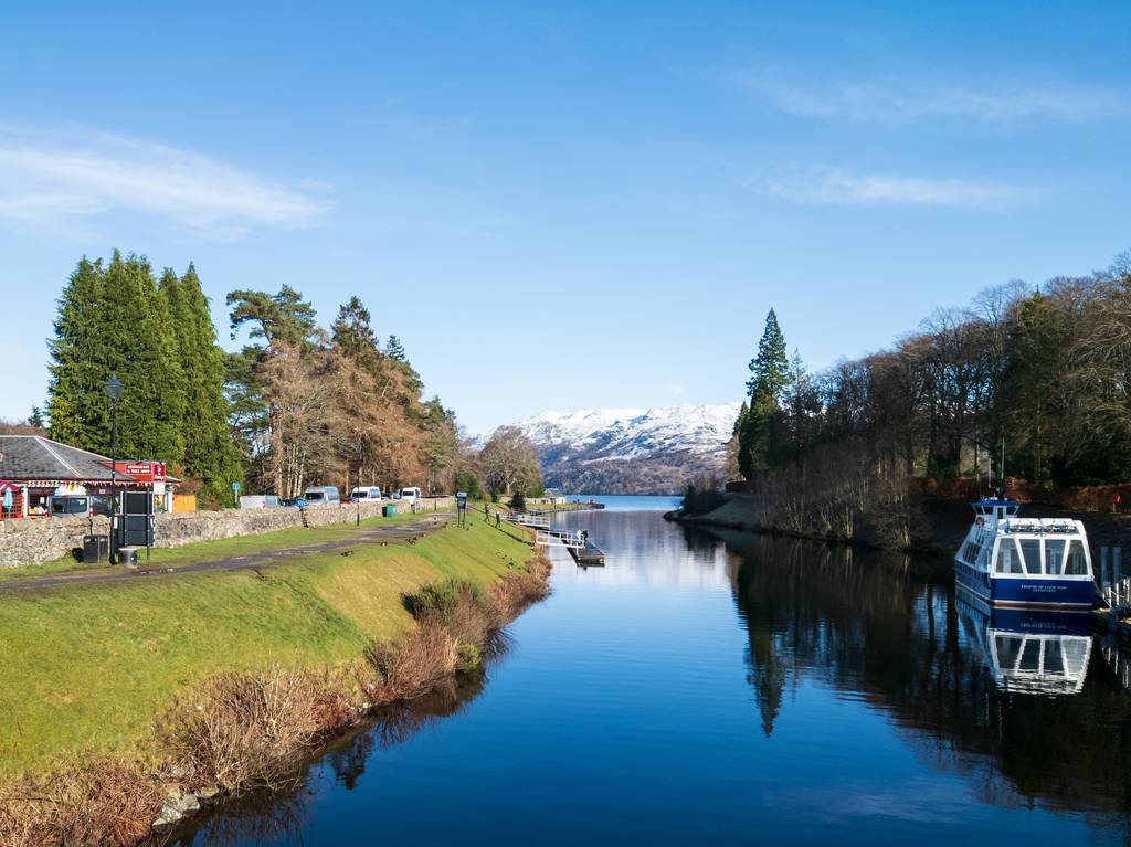 A serene Caledonian canal scene with a boat on the right, surrounded by lush greenery and trees under a clear blue sky, snow-capped mountains in the distance.