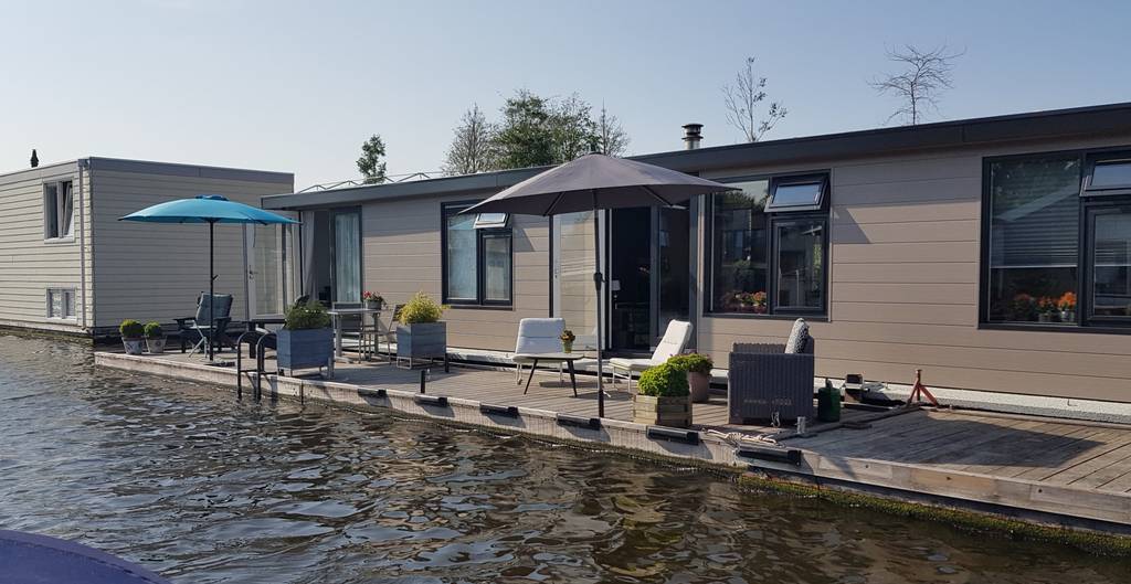 Modern houseboat on a calm canal with patio furniture, two umbrellas, and potted plants on the deck under a clear sky.