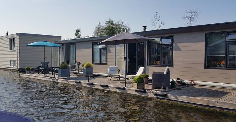 Modern houseboat on a calm canal with patio furniture, two umbrellas, and potted plants on the deck under a clear sky.