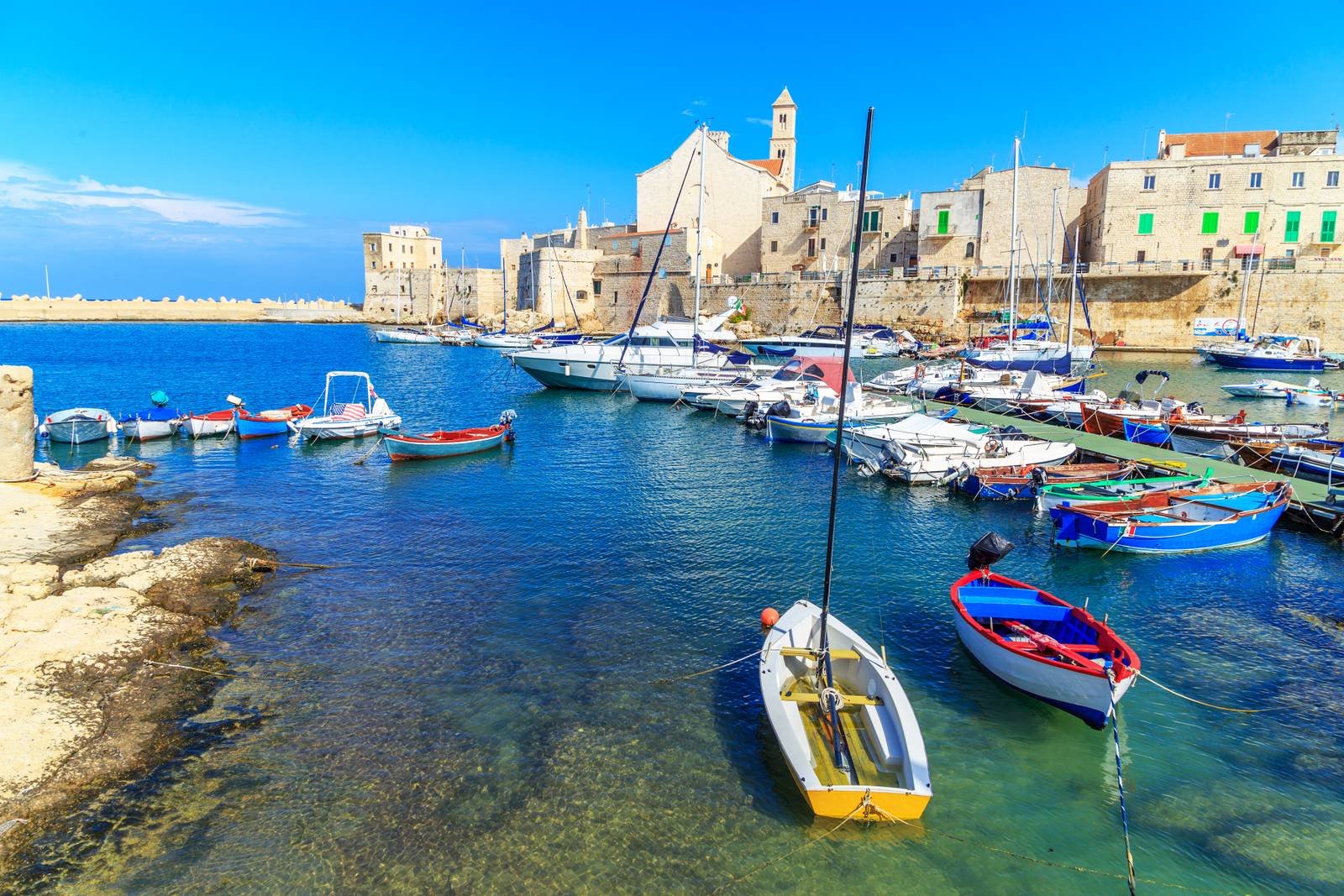 Colorful boats docked in the vibrant harbor of Giovinazzo with historic buildings under a clear blue sky and calm waters.