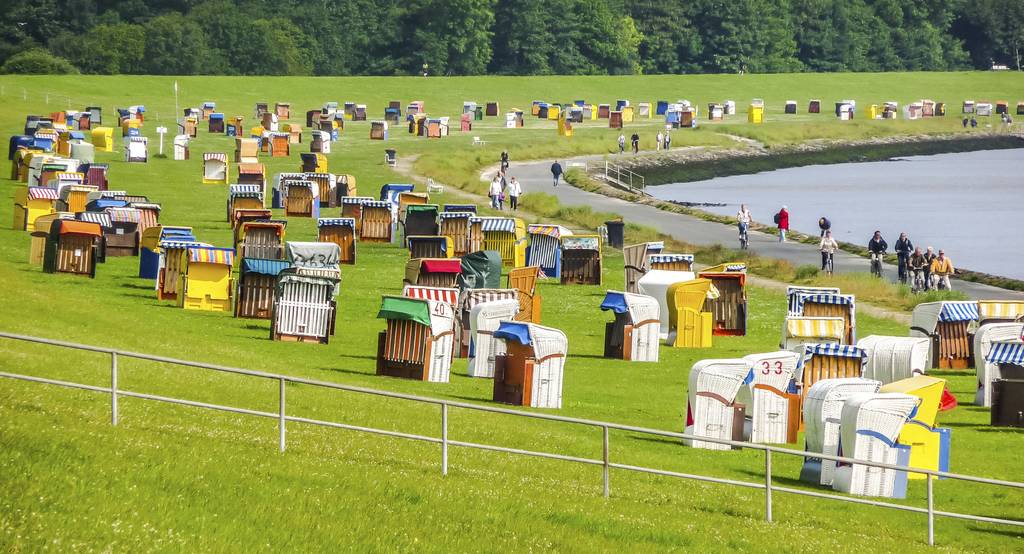 Grasbewachsener Park am See, übersät mit Dutzenden farbenfrohen gestreiften Strandkörben, mit Menschen, die auf einem Weg spazieren, und Bäumen im Hintergrund.