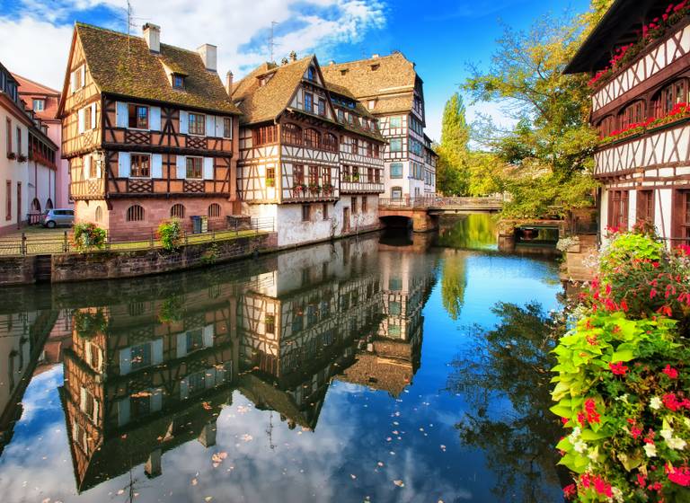 Charmante Fachwerkhäuser säumen einen ruhigen Kanal, in dessen Wasser sich leuchtende Blumen und der blaue Himmel spiegeln.