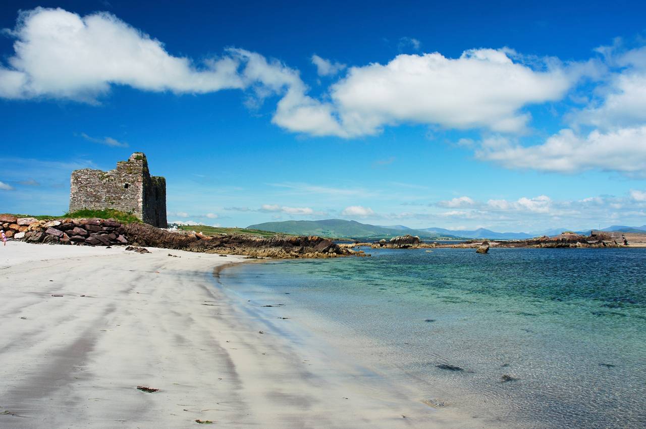A sandy beach with clear blue water and a stone ruin under a bright blue sky with scattered clouds. Hills are visible in the background.