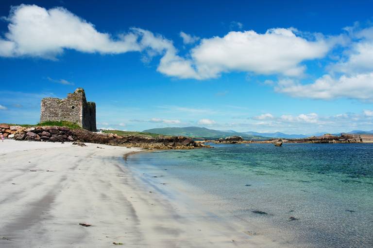 A sandy beach with clear blue water and a stone ruin under a bright blue sky with scattered clouds. Hills are visible in the background.