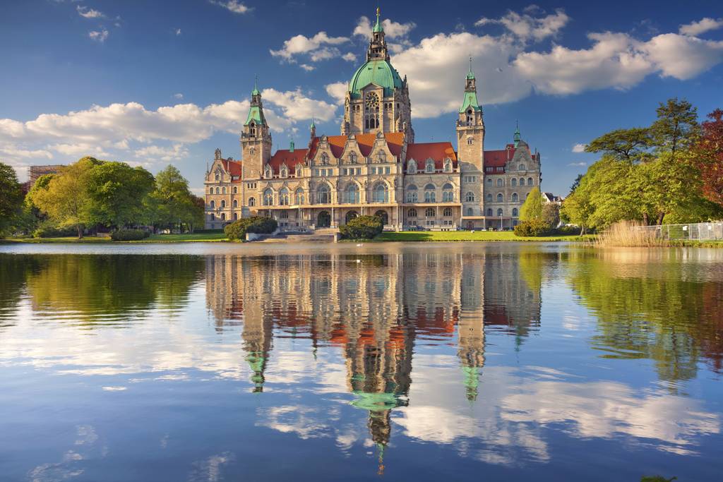 Majestätisches Gebäude mit grüner Kuppel und Türmen, spiegelt sich in einem stillen See unter einem teils bewölkten blauen Himmel