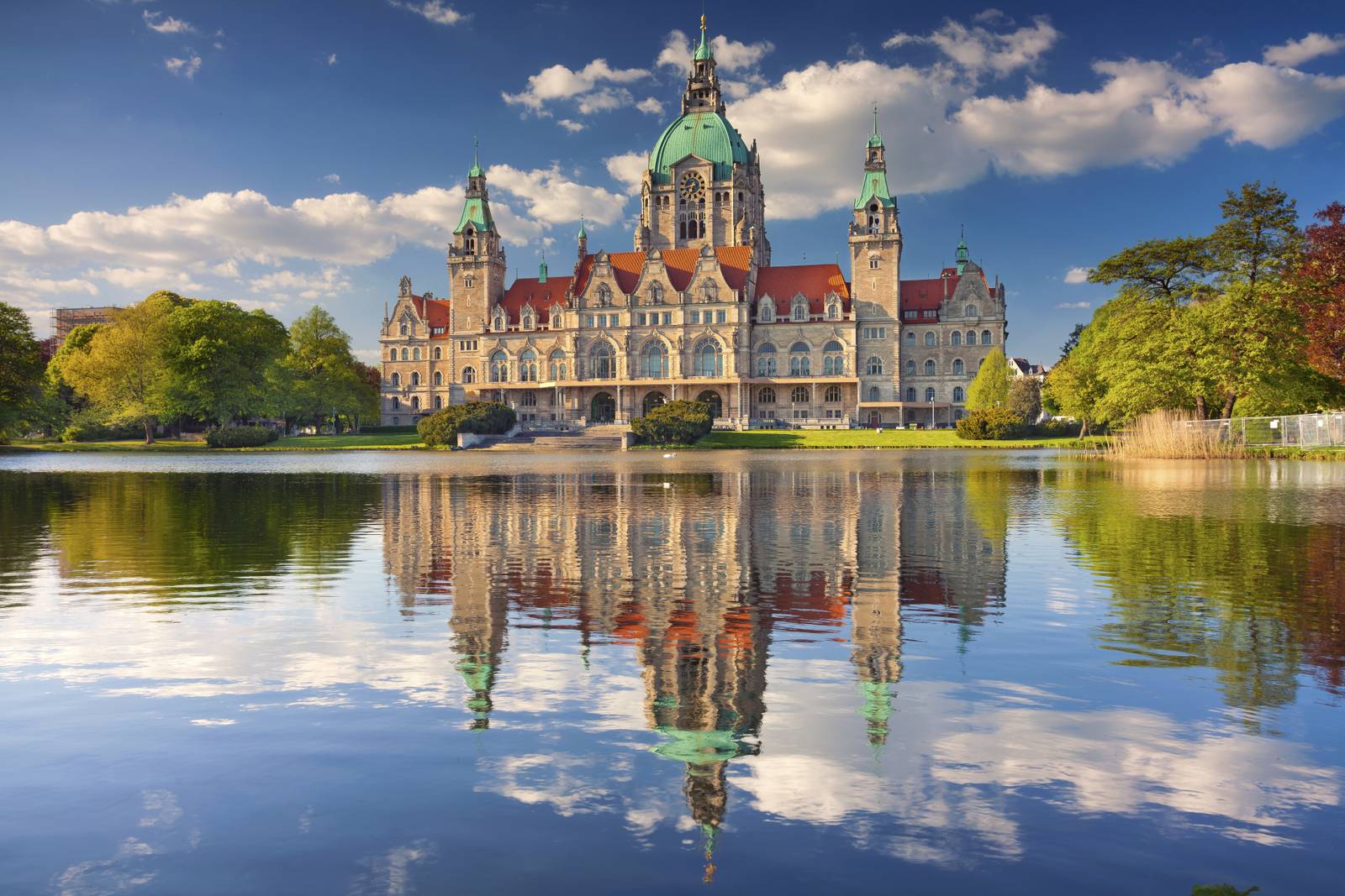 Majestätisches Gebäude mit grüner Kuppel und Türmen, spiegelt sich in einem stillen See unter einem teils bewölkten blauen Himmel