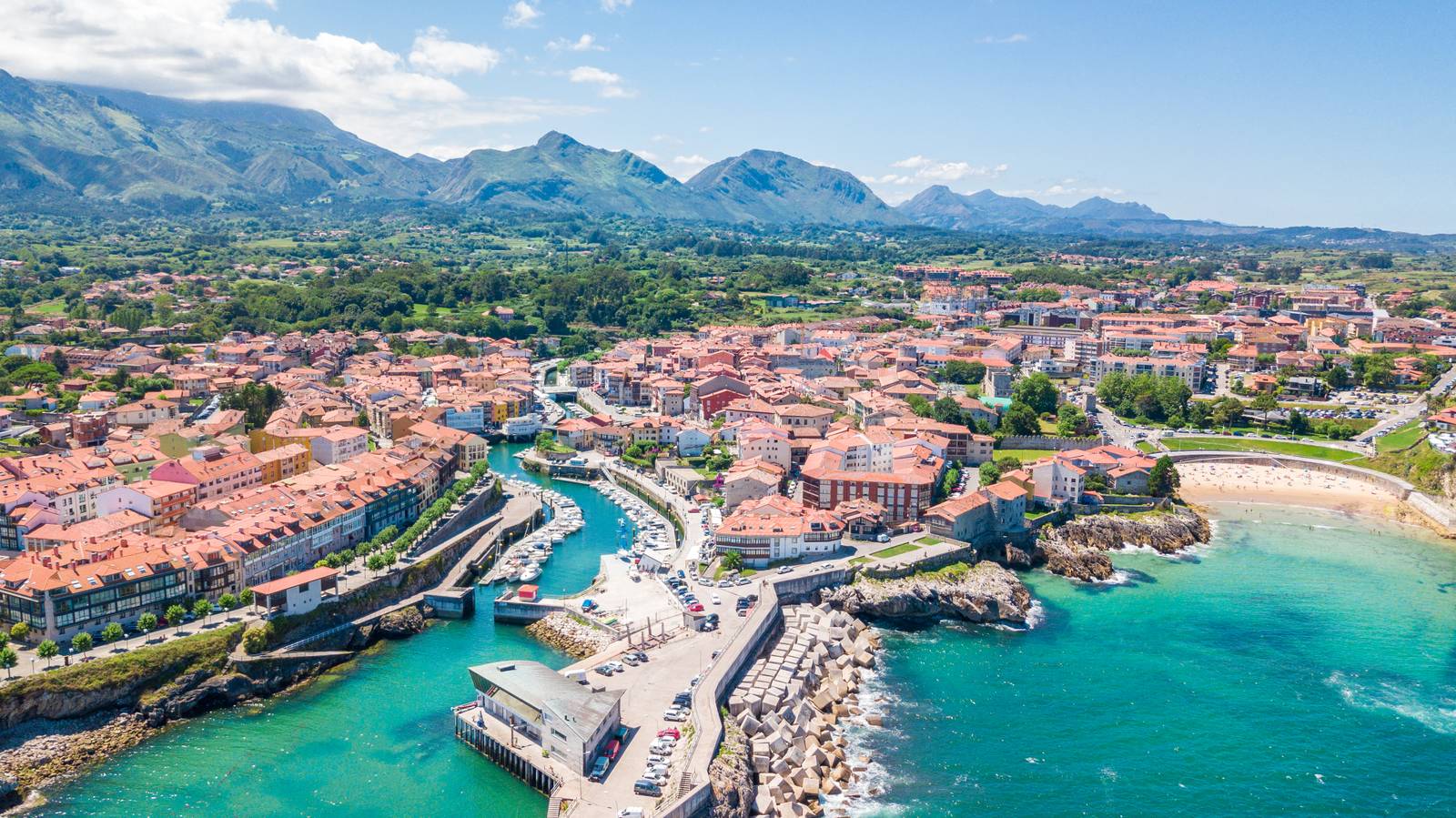 Aerial view of Llanes town with red-roofed buildings, a marina, and green mountains in the background under a clear blue sky.