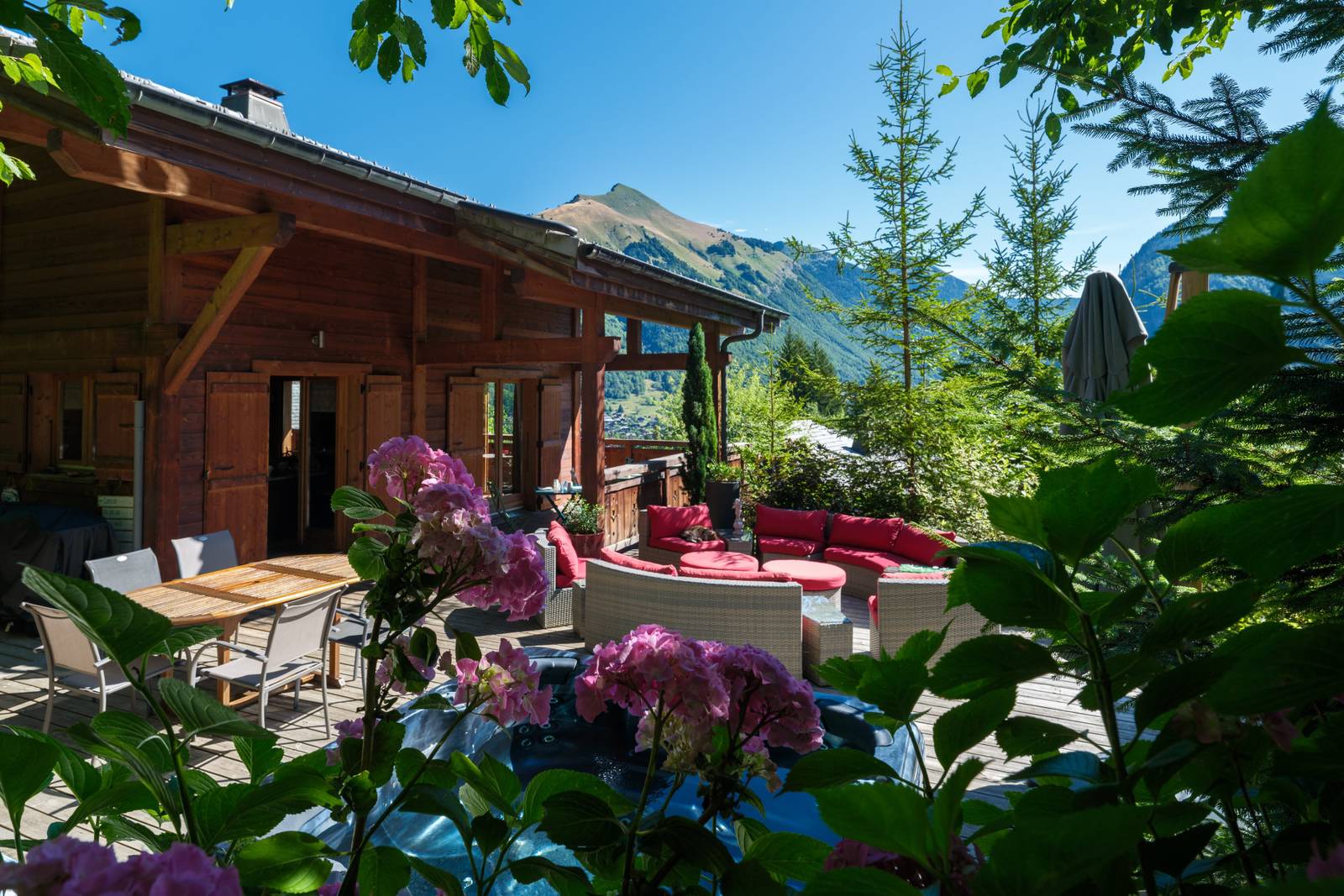 Bergchalet-Terrasse mit Holzmöbeln und rosa Blumen, umgeben von Bäumen. Malerische Berge im Hintergrund unter klarem blauem Himmel.
