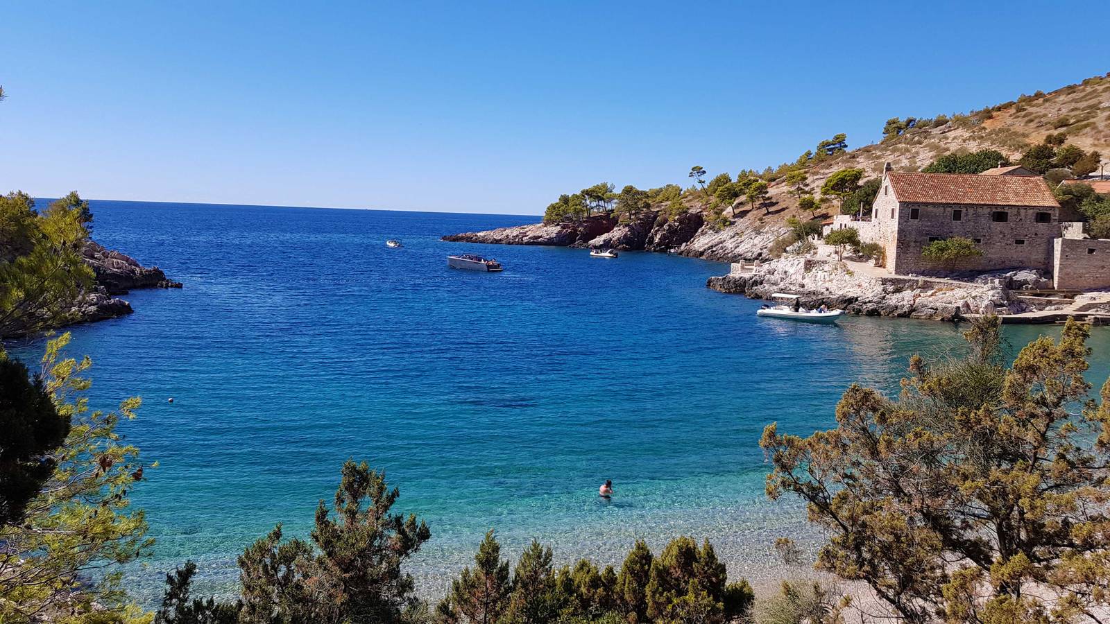 Coastal scene with clear blue water, rocky shoreline, and a stone building. Trees and shrubs frame the view under a clear blue sky.