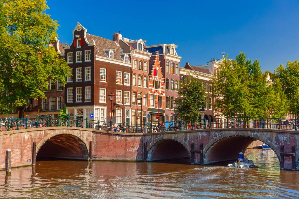 Historische Gebäude und eine Brücke über einen Kanal in Amsterdam, mit klarem blauem Himmel und grünen Bäumen, die sich im Wasser spiegeln.