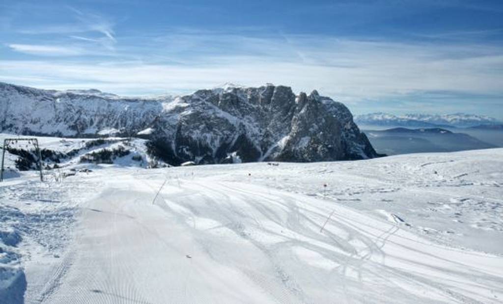 Verschneite Skipiste mit Skispuren, die auf zerklüftete Berggipfel unter einem strahlend blauen Himmel zuführen.