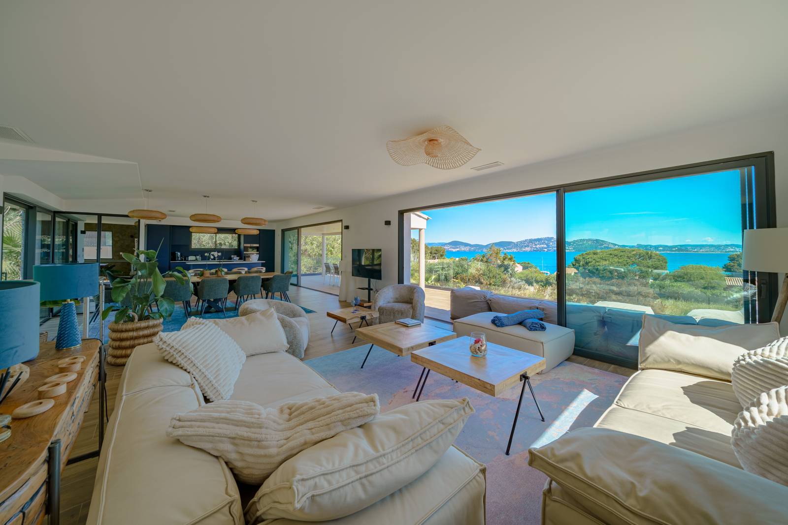 Geräumiges Wohnzimmer mit weißen Sofas, Holztischen, großen Fenstern und Meerblick; im Hintergrund ein offener Küchenbereich.
