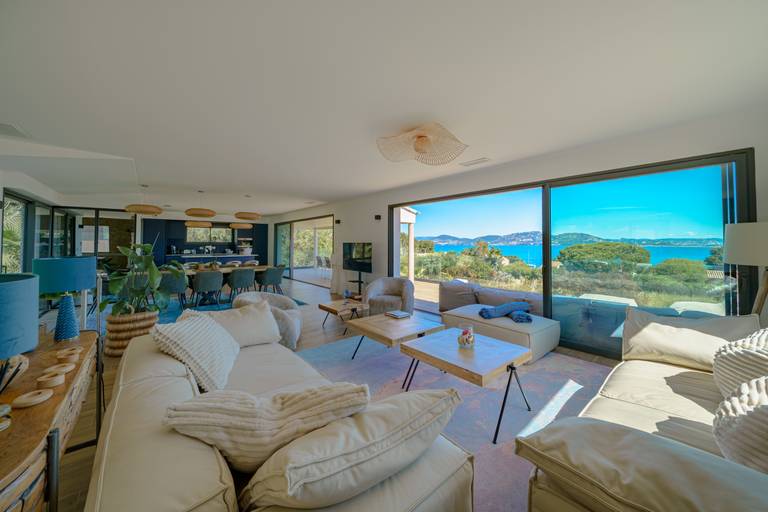 Geräumiges Wohnzimmer mit weißen Sofas, Holztischen, großen Fenstern und Meerblick; im Hintergrund ein offener Küchenbereich.