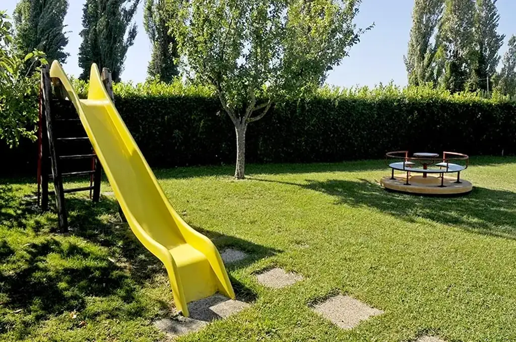 A sunny playground with a yellow slide, a merry-go-round, a tree, and a green hedge surrounding the grassy area.