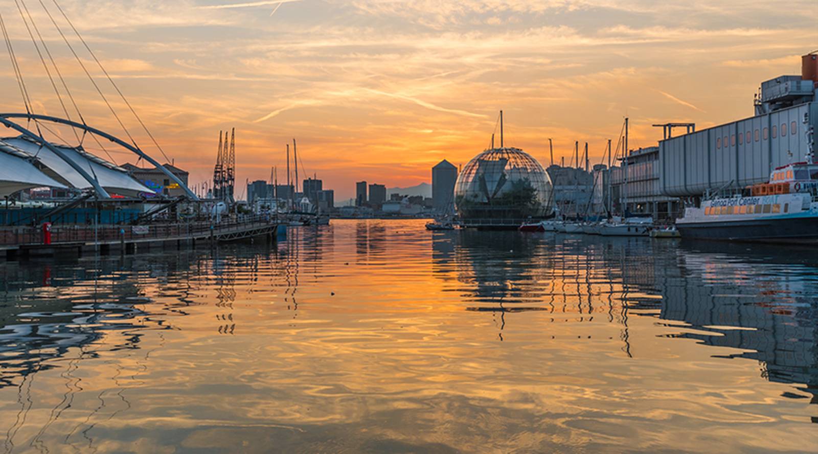 Sonnenuntergang über einem Hafen mit ruhigem Wasser, das den orangefarbenen Himmel widerspiegelt, festgemachten Booten und einer markanten Glaskuppel im Hintergrund