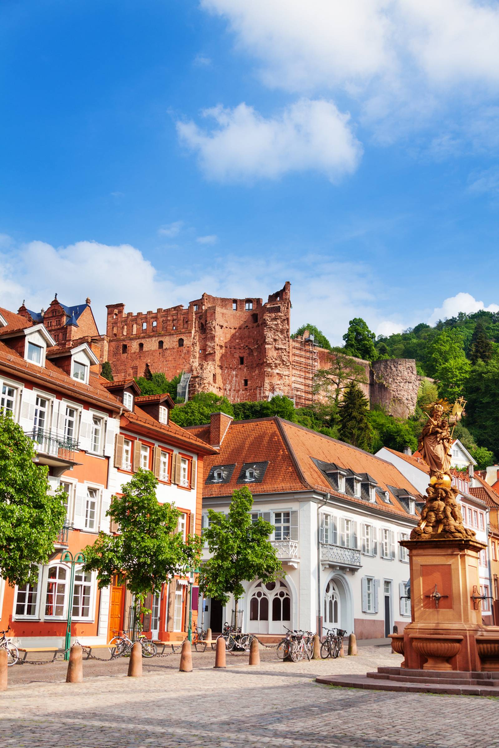 Das Heidelberger Schloss auf einem Hügel, mit Blick auf bunte historische Gebäude und eine Statue auf einem sonnigen Platz unter blauem Himmel