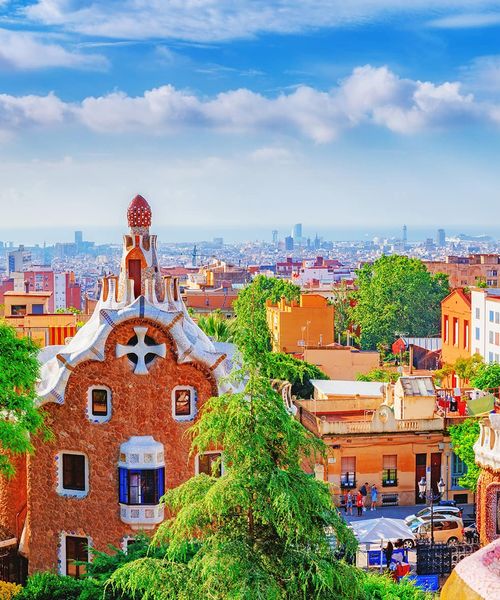 A view overlooking Park Guell which is a vibrant outdoor collection of gardens and architecture located in Barcelona. A red brick building with a white cross and white trim details is in the foreground with several trees. The city of Barcelona can be seen in the background with clouds scattering a blue sky.