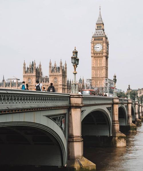 A long green bridge with a bus and people traveling on top of it and many tall ornate gold buildings in the background