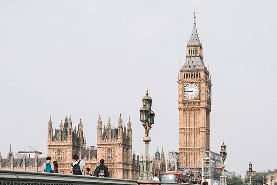 A long green bridge with a bus and people traveling on top of it and many tall ornate gold buildings in the background