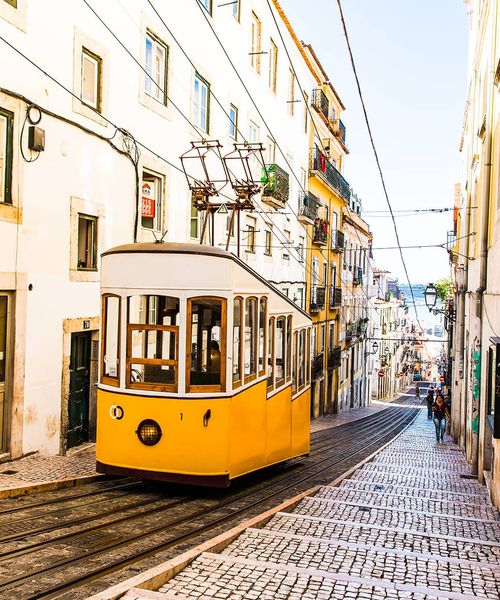 A white and yellow trolley traveling up a hill between two buildings with many wires crossing above the trolley
