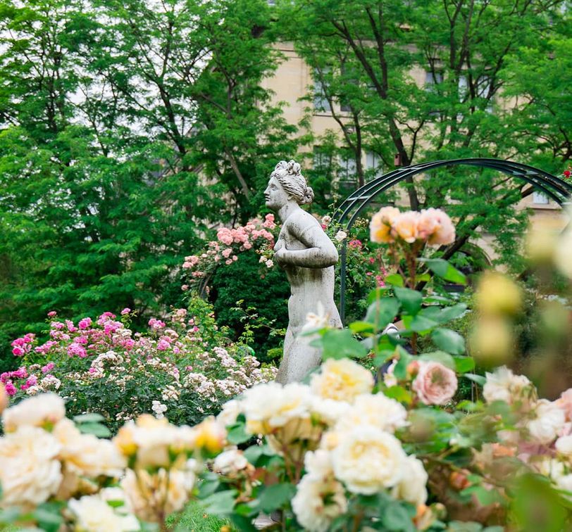 A stone figure of a woman surrounded by many pink and white roses and lush greenery