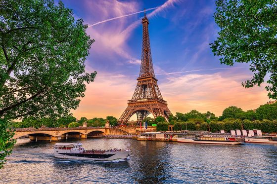 The Eiffel Tower with a vibrant sunset in the background and a body of water in the foreground with a boat passing in front